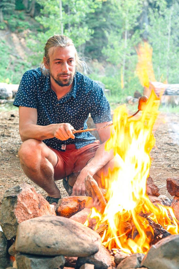 Michael crouched next to a campfire roasting a hotdog on a stick