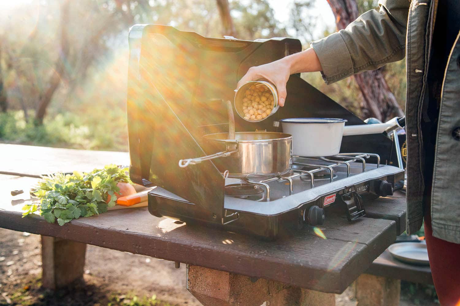 Woman pouring a can of chickpeas into a pot on a camping stove.