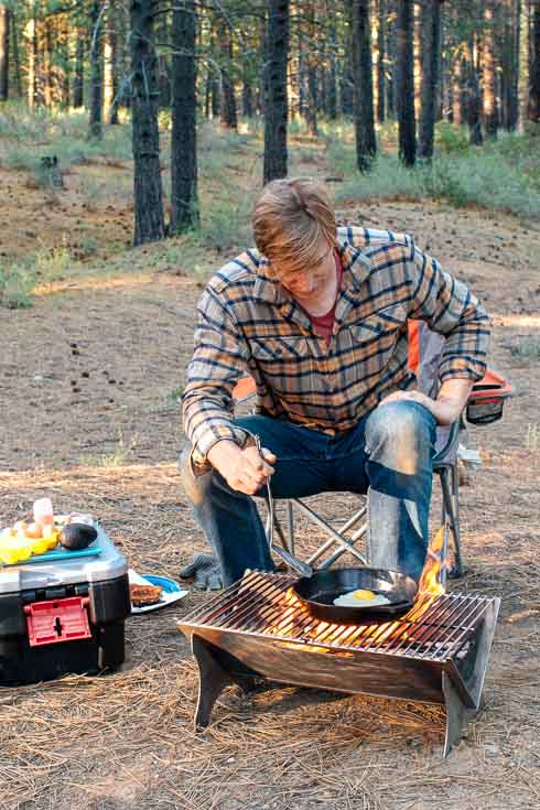 Man sitting next to a campfire cooking an egg in a cast iron skillet