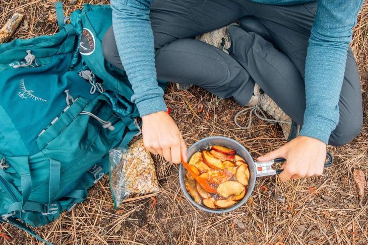 Megan sitting on the ground cooking an apple crisp in a backpacking pot.