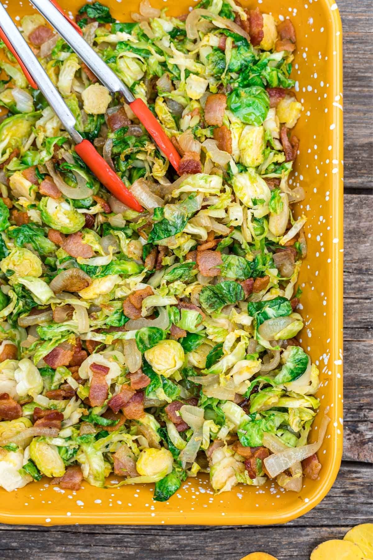 Shredded brussels sprouts in a yellow dish with red tongs