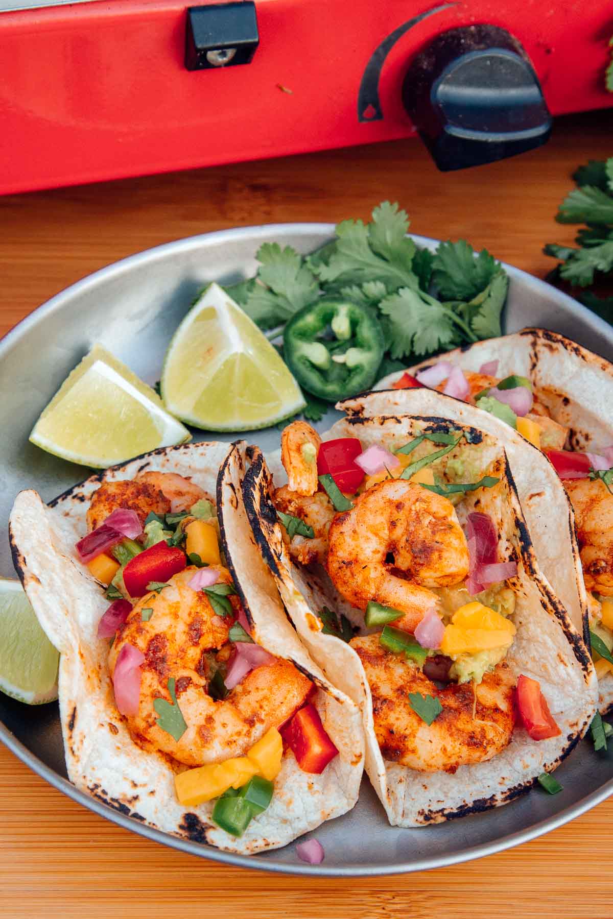 Three shrimp tacos on a silver plate with sliced limes and jalapenos on the side.