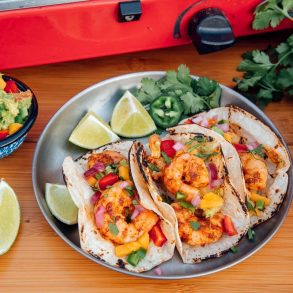 Three shrimp tacos on a silver plate with sliced limes and a small bowl of guacamole on the side.