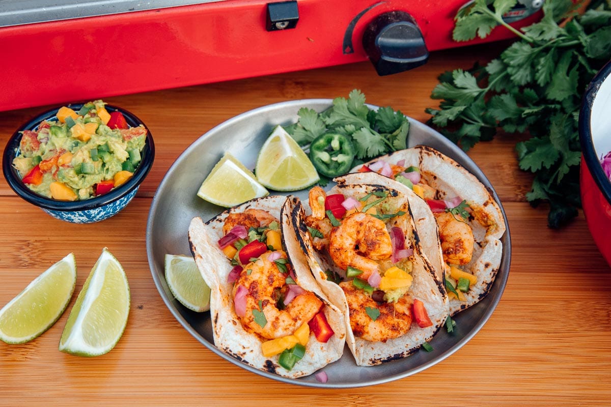 Three shrimp tacos on a silver plate with sliced limes and a small bowl of guacamole on the side.