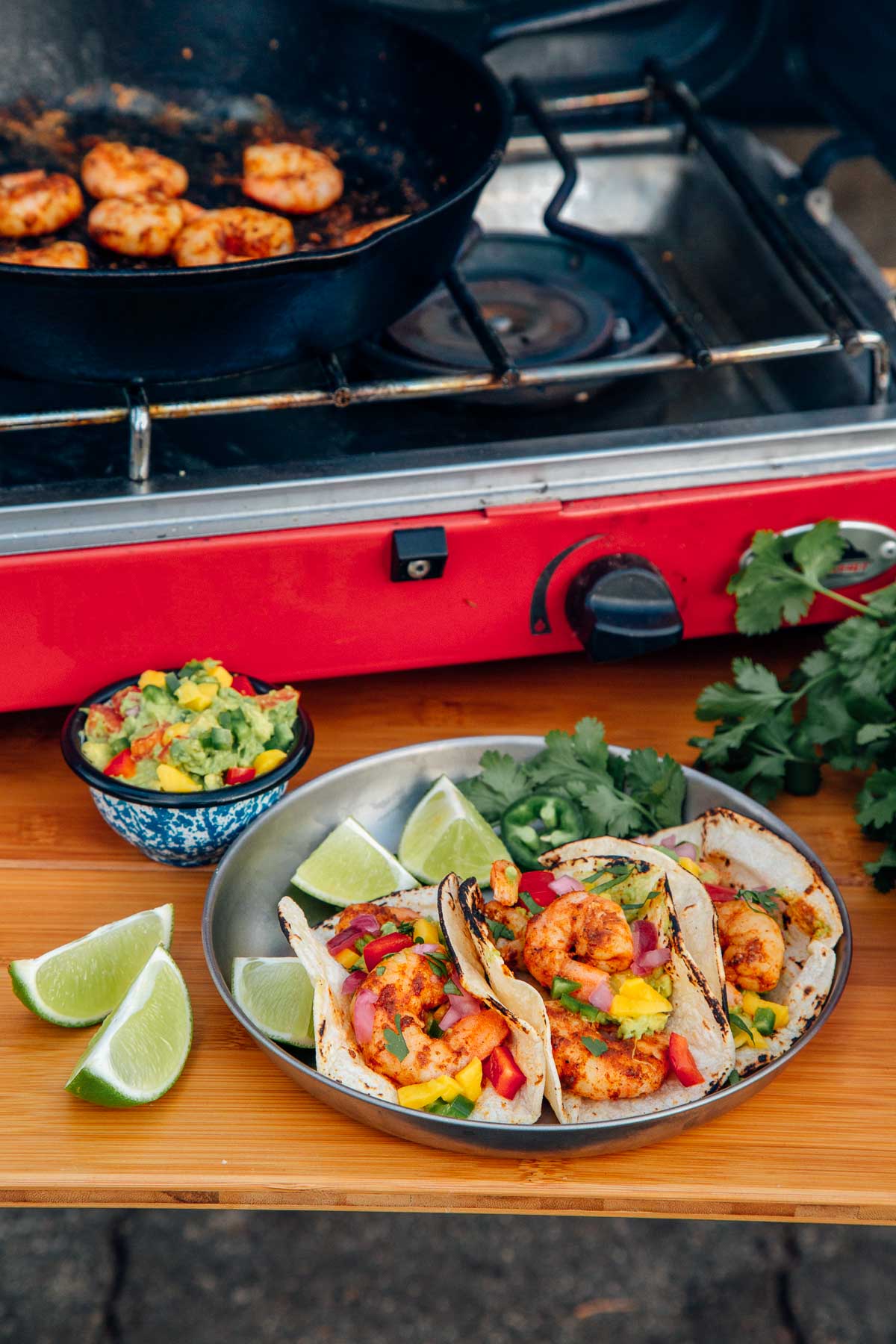 Three shrimp tacos on a silver plate with sliced limes and a small bowl of guacamole on the side. A camp stove is in the background.