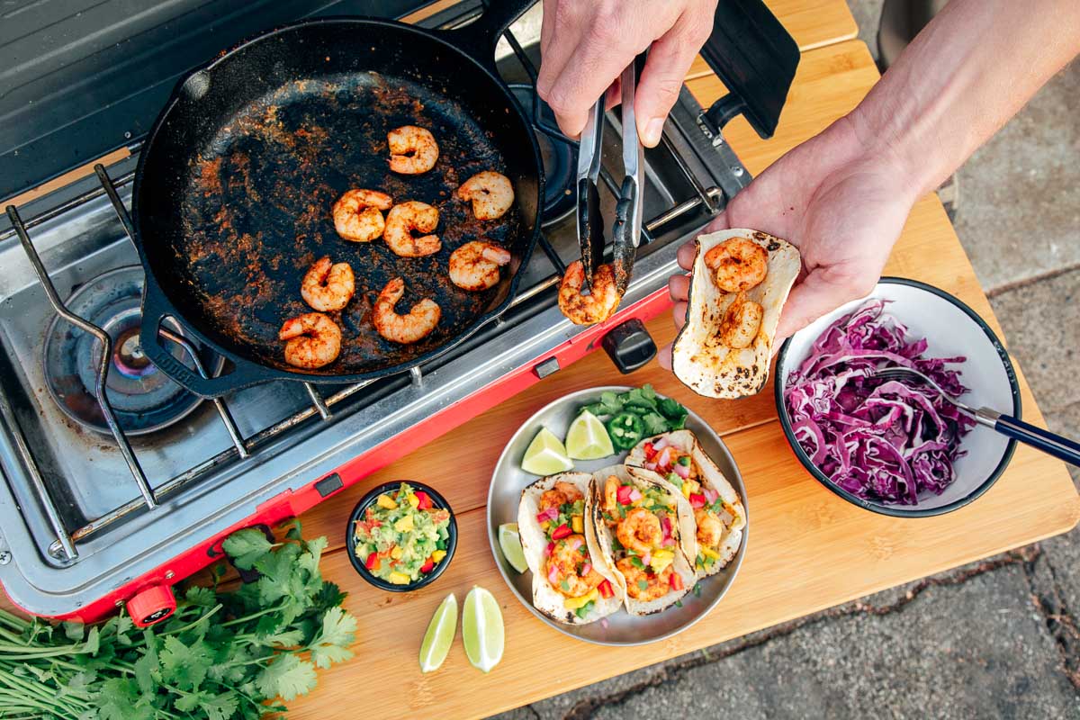 Michael building a shrimp tacos. A cast iron skillet of shrimp sits on a camp stove in the background.