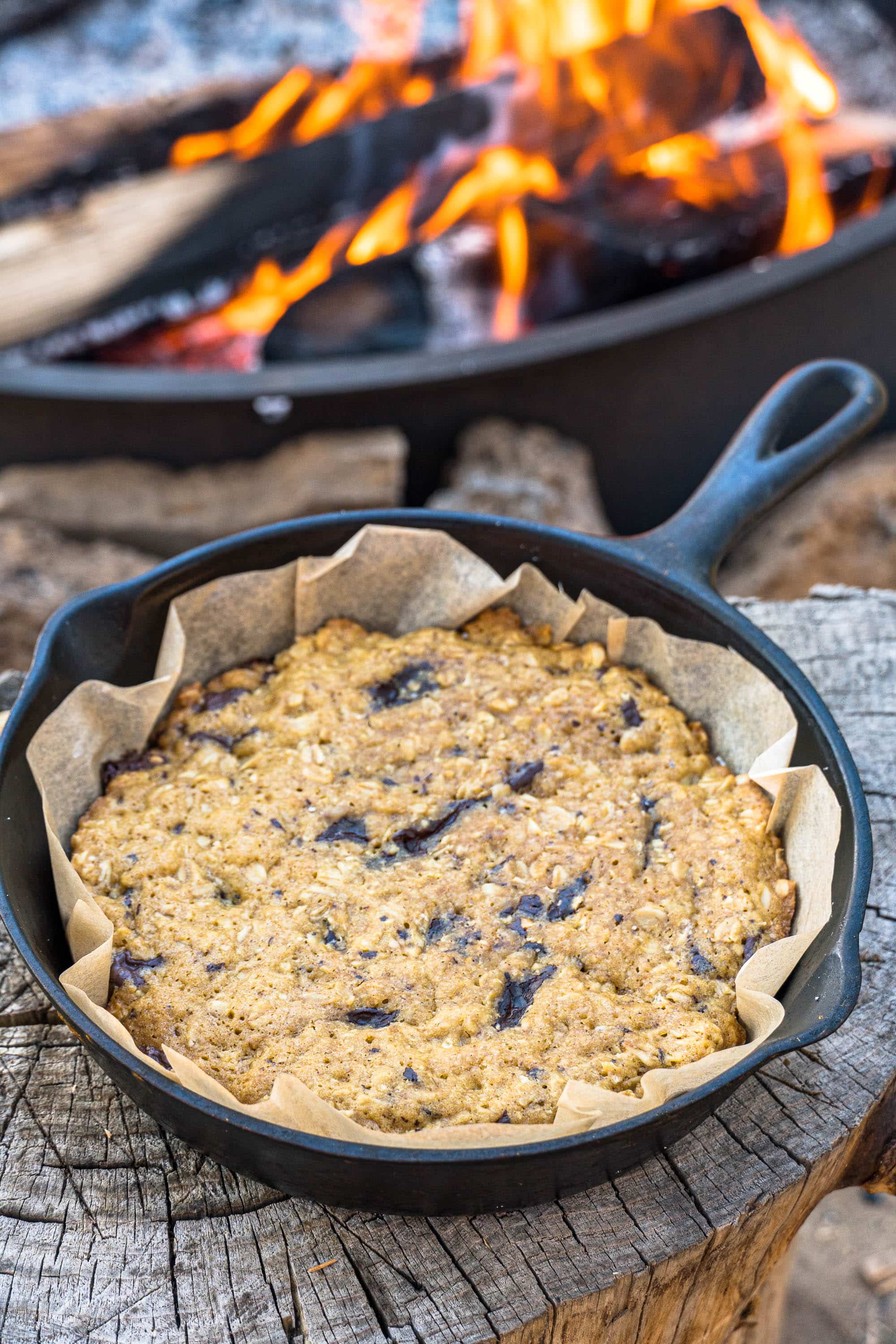 A chocolate chip cookie in a cast iron skillet set on a wood log.