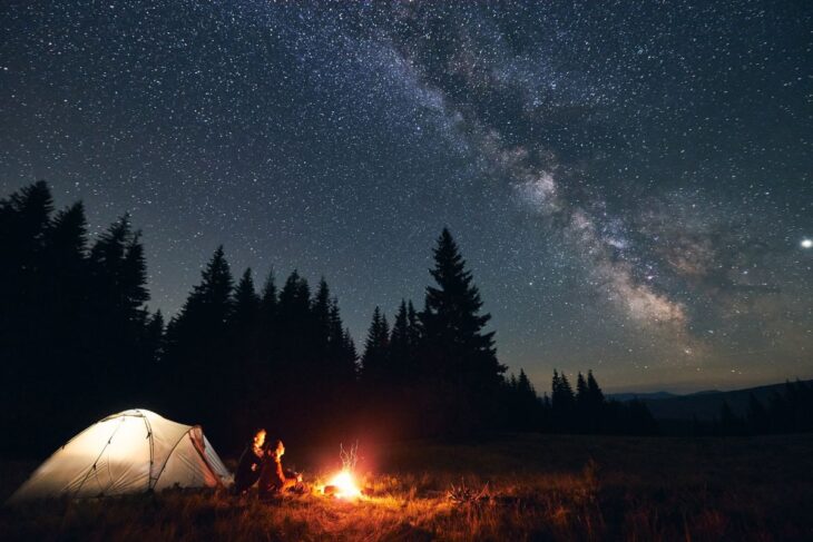 A couple sits next to a tent and a campfire under a starry sky