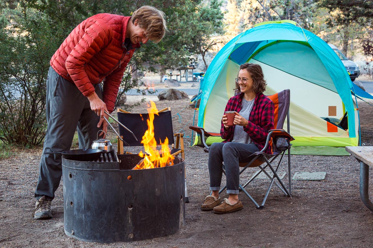 Megan and Michael around a campfire. Michael is using tongs to move something and Megan is sitting with a cup of coffee in hand.