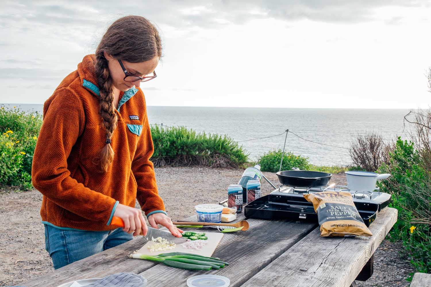 Woman chopping jalapeÃ±os at a campsite. A camping stove and other nacho ingredients are visible in the background.