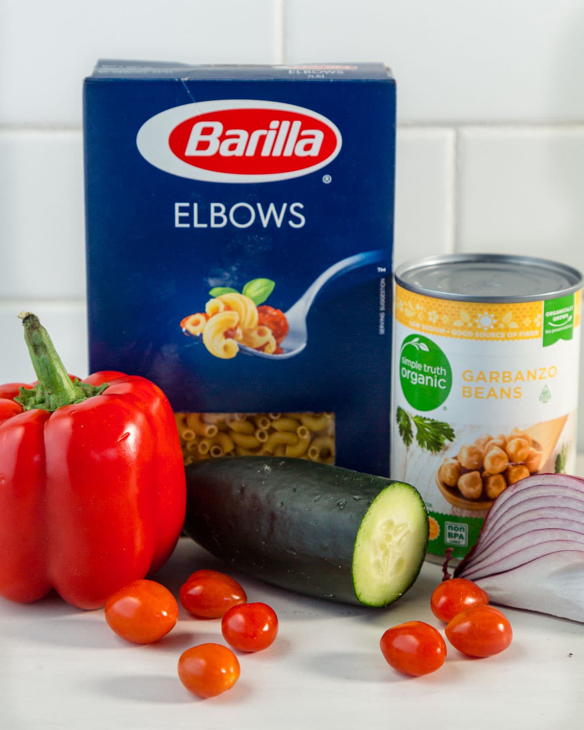 Ingredients for pasta salad: box of pasta, red bell pepper, can of chickpeas, cucumber, tomatoes, and red onion slice.