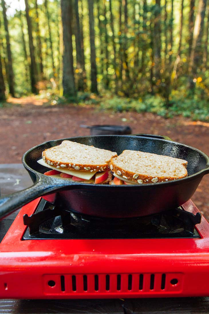 Grilled cheese sandwiches in a skillet on a camp stove with forest in the background