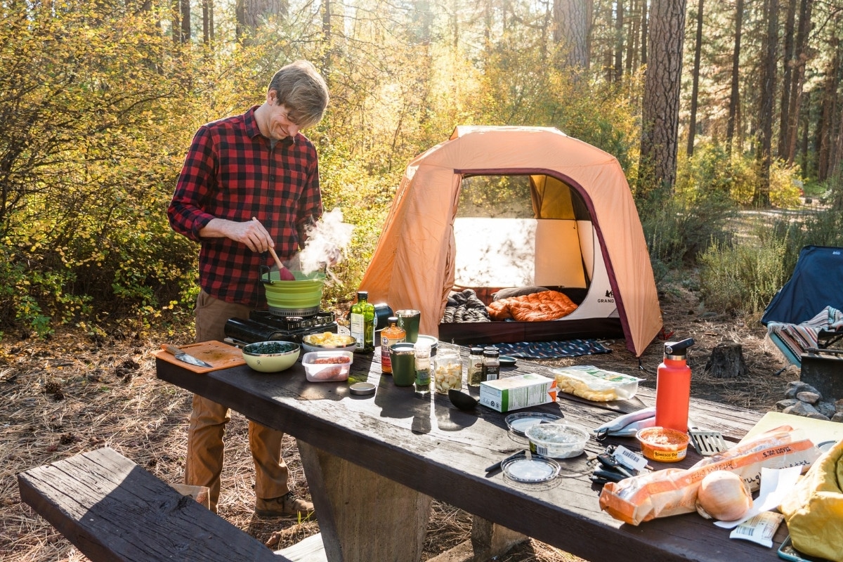 Michael standing at a camping table stirring tortellini soup in a pot. A camp scene is in the background