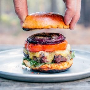 A man placing a bun on top of a grilled burger stacked with onions, tomatoes, and lettuce