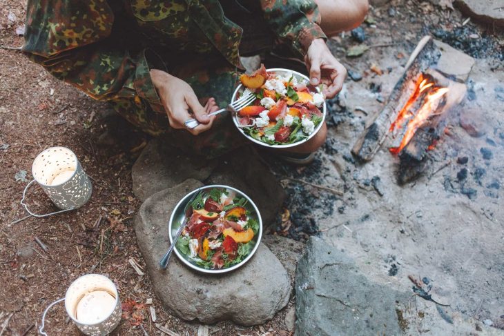 Person holding a plate of peach caprese salad next to a campfire