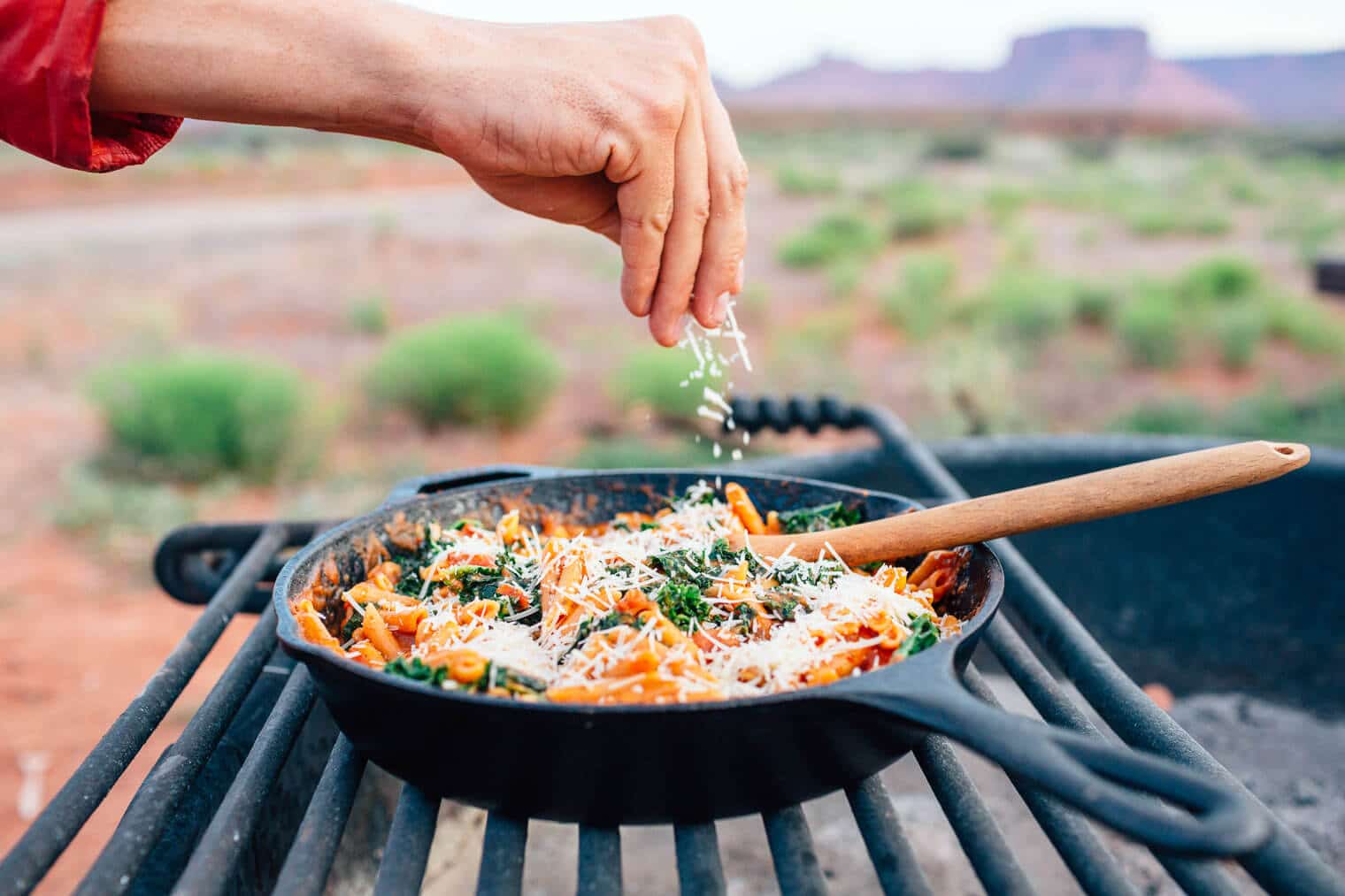 Michael sprinkling cheese into a skillet of pasta that is sitting on a campfire