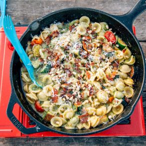 Overhead shot of pesto pasta in a cast iron skillet on a camping stove topped with cheese and pine nuts.