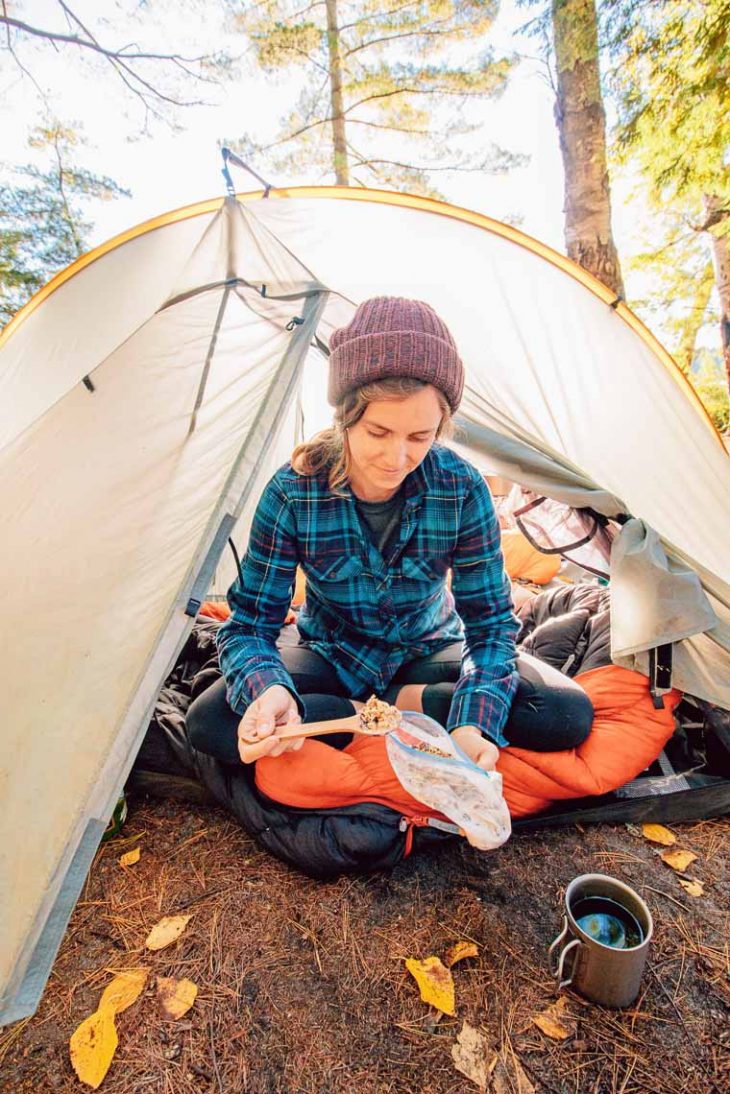 Megan sitting at the entrance of a backpacking tent eating granola out of a baggie