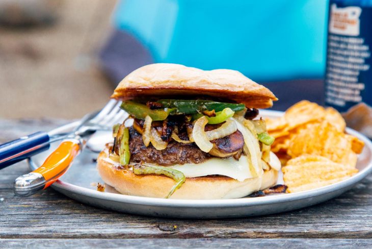 A burger topped with green peppers and onions on a blue plate and camping scene in the background