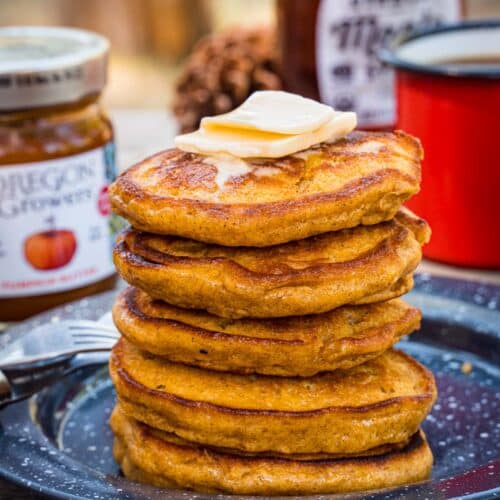 Pumpkin pancakes stacked on a plate. A coffee cup and a bottle of maple syrup are in the background.