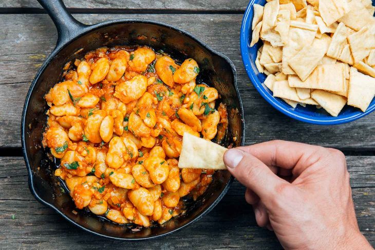 Hand holding a pita chip with Spanish White Bean Dip in a cast iron skillet