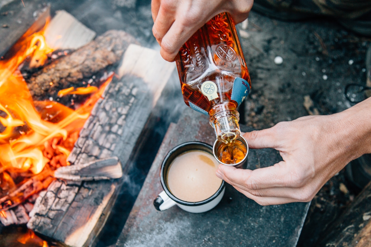 Pouring whiskey into a mug of pumpkin spiced chai with a campfire in the background