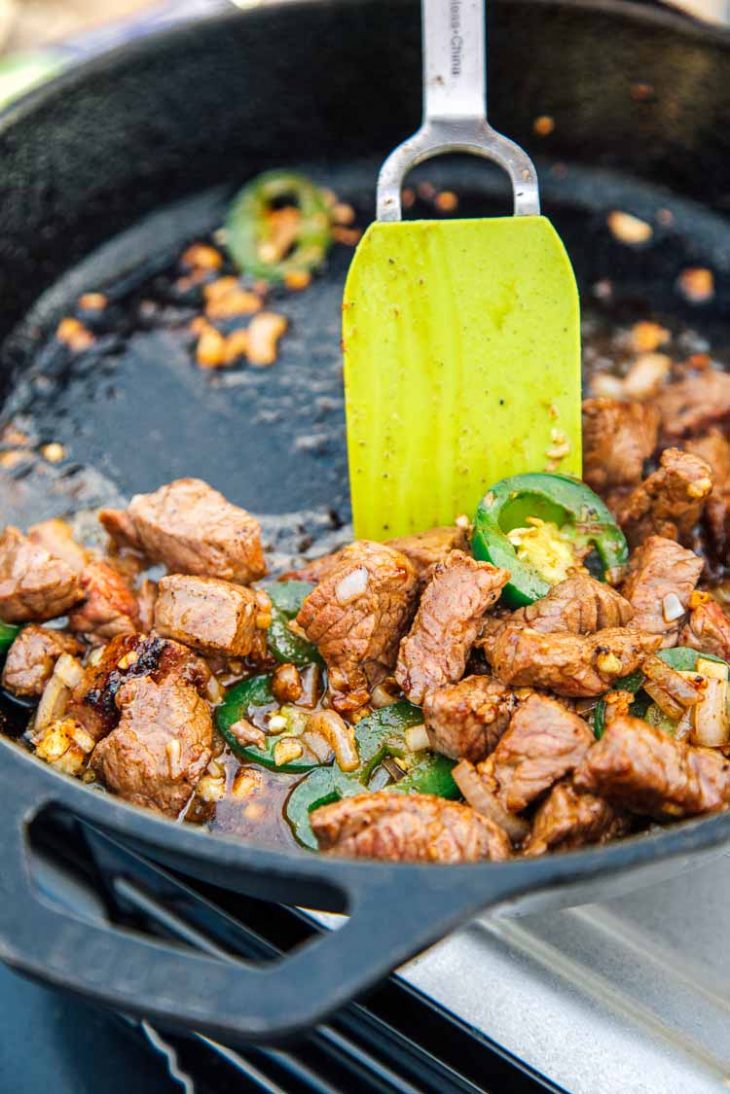 Action shot of a spatula satueeing steak, jalapeños, garlic, and shallots in a cast iron skillet.