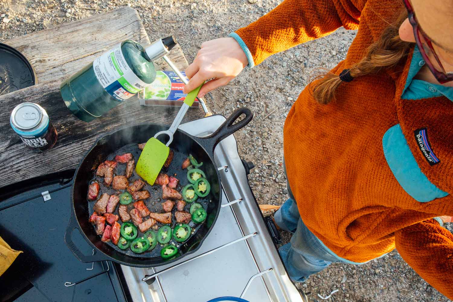 Overhead shot of a woman cooking steak and jalapeños in a cast iron skillet over a camping stove.