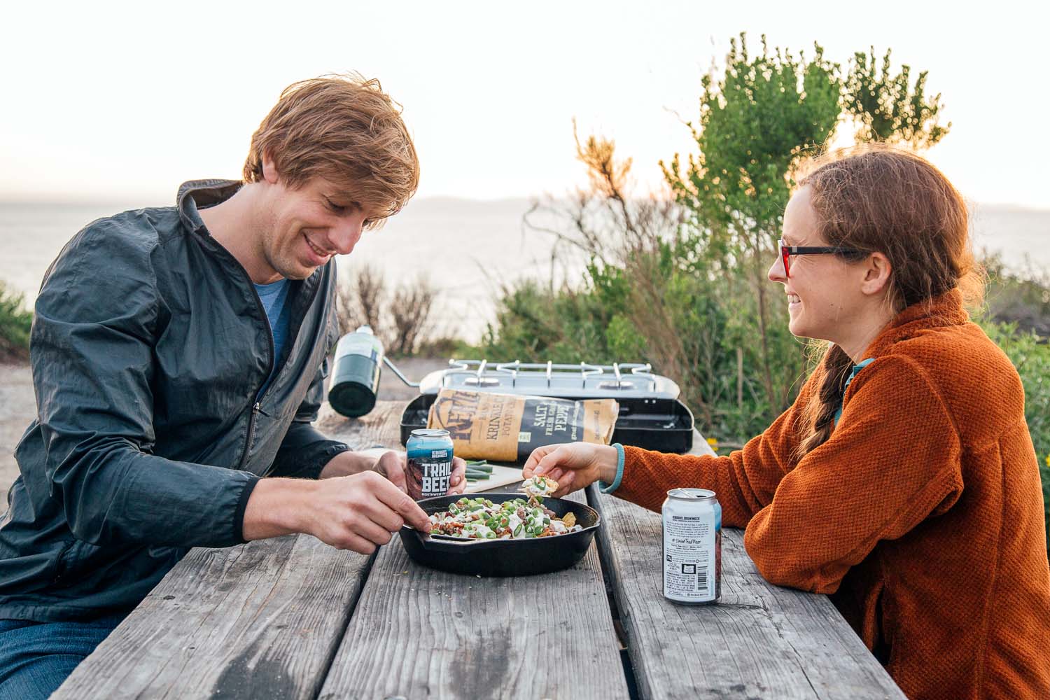 Man and woman sitting at a picnic table sharing a plate of nachos at a camp site with bushes in the background.