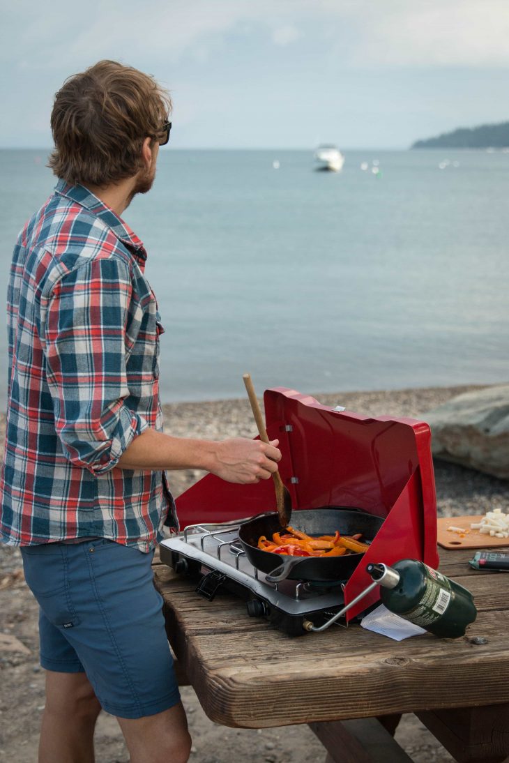 A man cooking stirfry udon noodles in a pan on a camp stove with water in the background