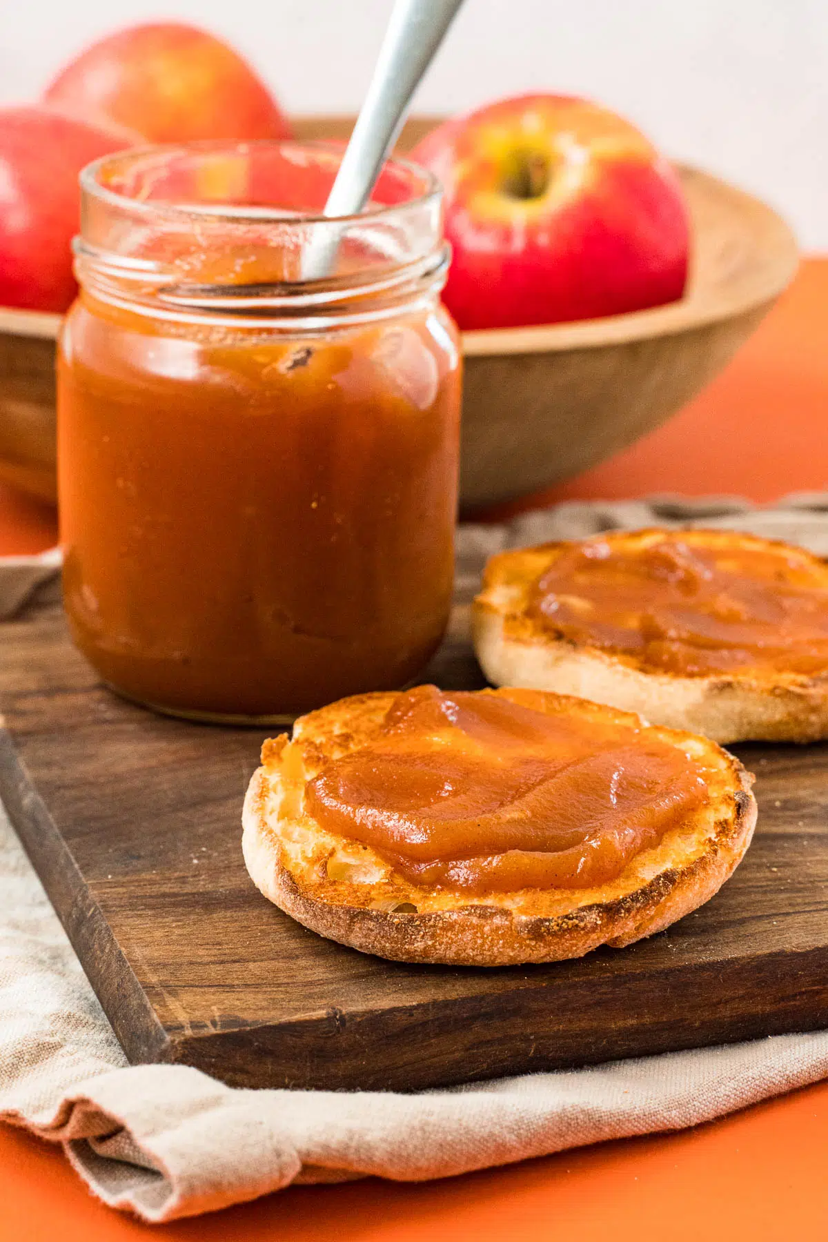 A jar of apple butter next to an english muffin topped with apple butter.