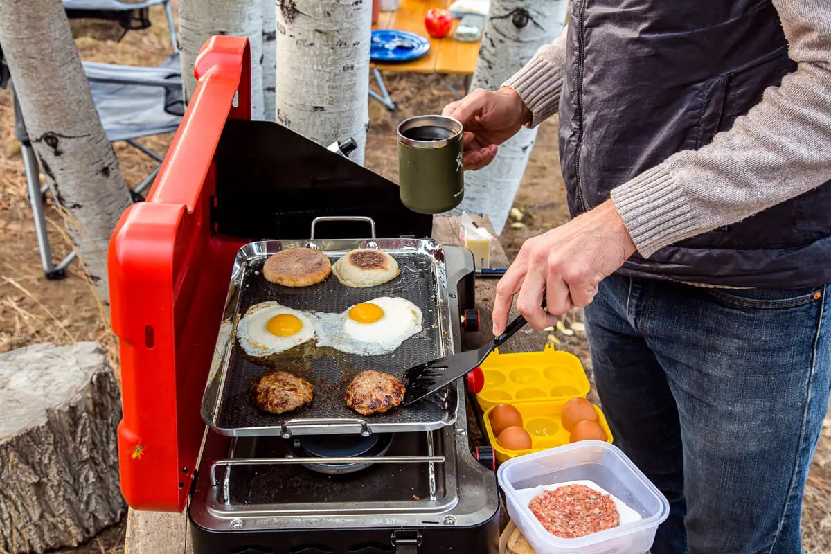 Michael is holding a mug of coffee in one hand and is holding a spatula to flip a sausage on a griddle with the other hand.