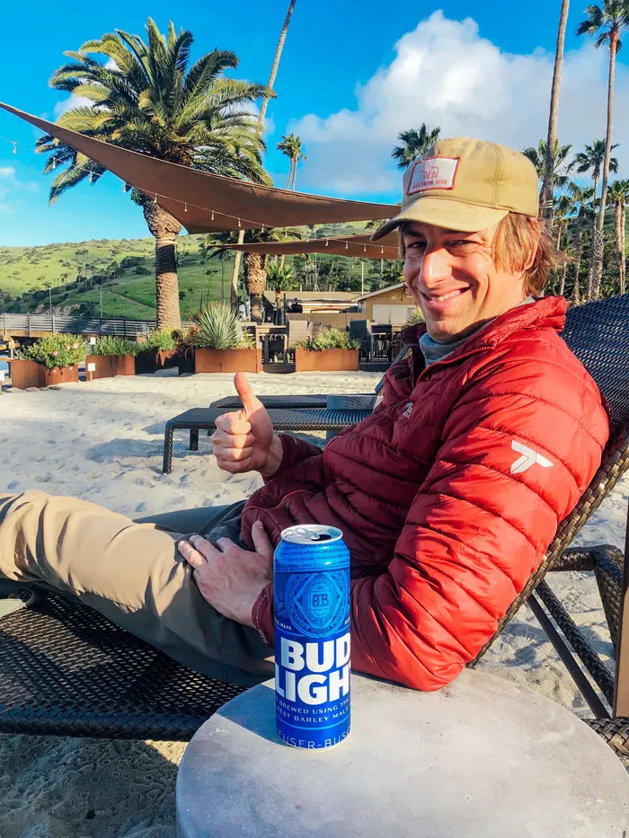 Man on a beach with a beer giving a thumbs up.