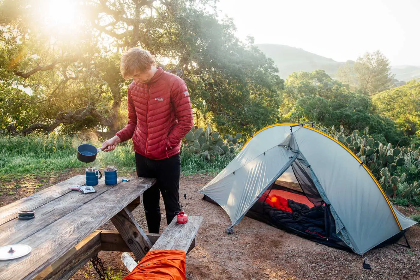 Man making camp coffee at a picnic table with a tent in the background