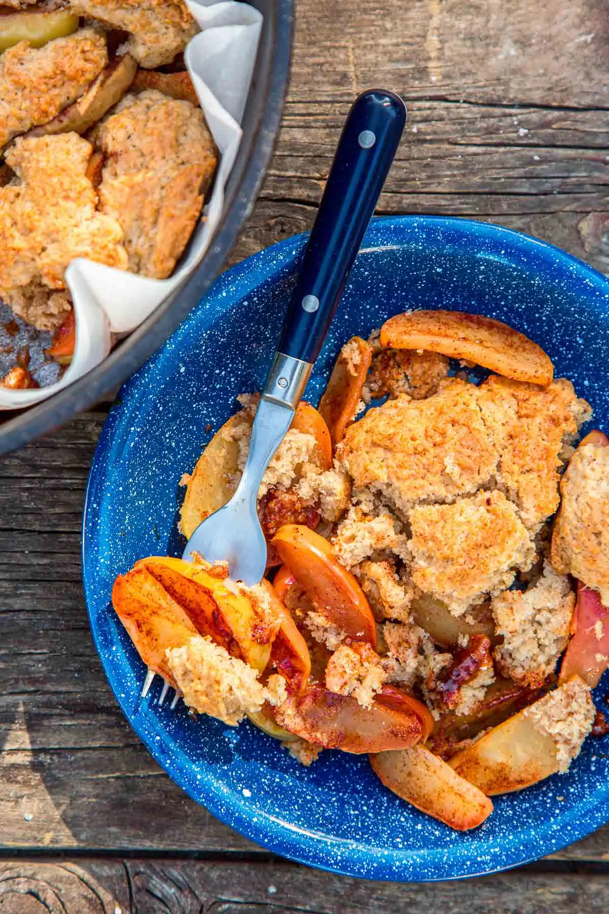 Apple cobbler in a blue bowl with a fork next to a Dutch oven