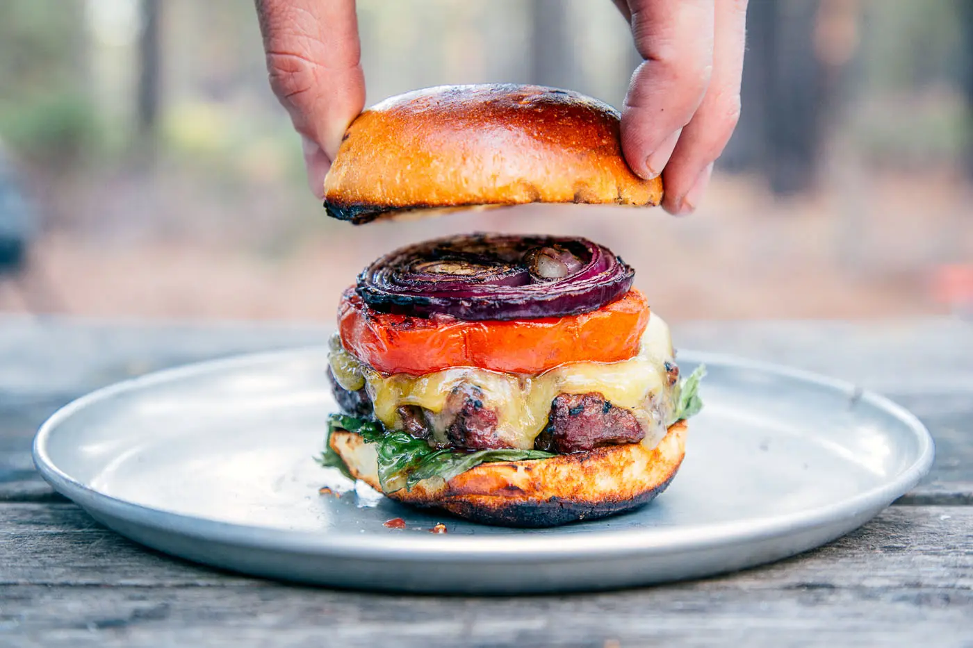 A man placing a bun on top of a grilled burger stacked with onions, tomatoes, and lettuce