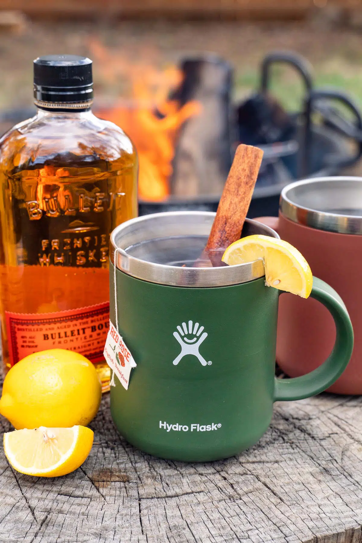 A mug of hot toddy with ingredients and a campfire in the background