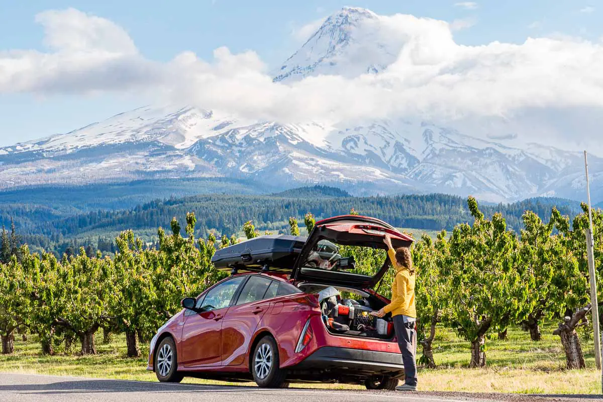 Megan opening the trunk of a red car which is parked on the side of the road next to an orchard. Mt. Hood, which is shrouded with clouds, can be seen in the distance.