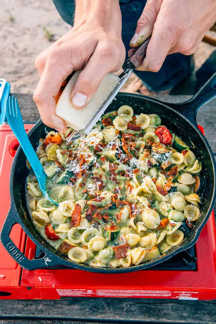 Man grating parmesan cheese over a skillet of pesto pasta