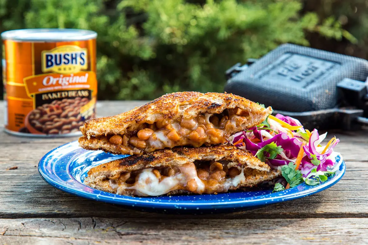 A sandwich filled with beans and cheese cut diagonally in half, resting on a blue plate with a pie iron in the background