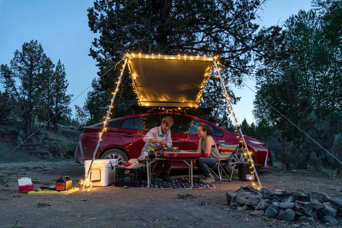 Megan and Michael sit next to a red car that has an awning set up with string lights.