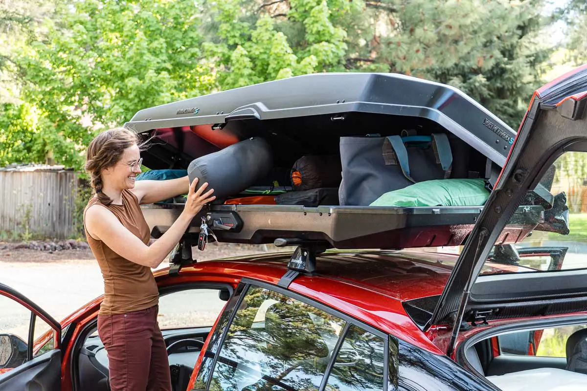 Megan stands next to a car with a roof box. She is placing a sleeping bag into the box