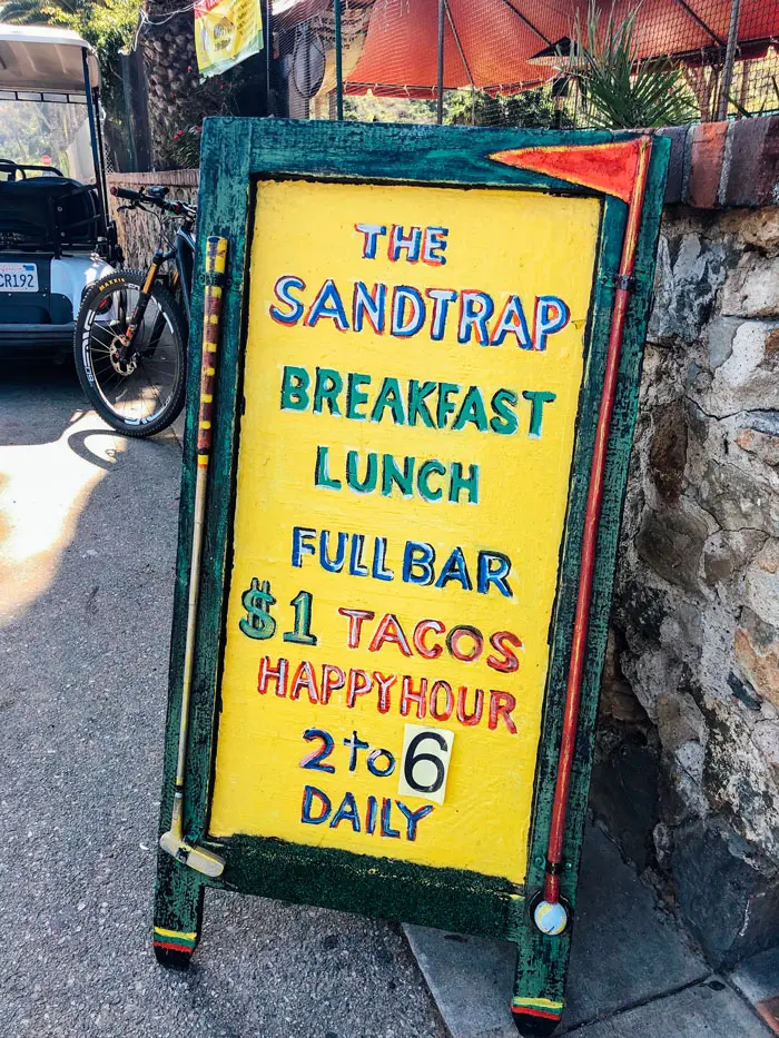 Painted yellow sign that reads "The Sandtrap. Breakfast, lunch, full bar. $1 Tacos Happy Hour"