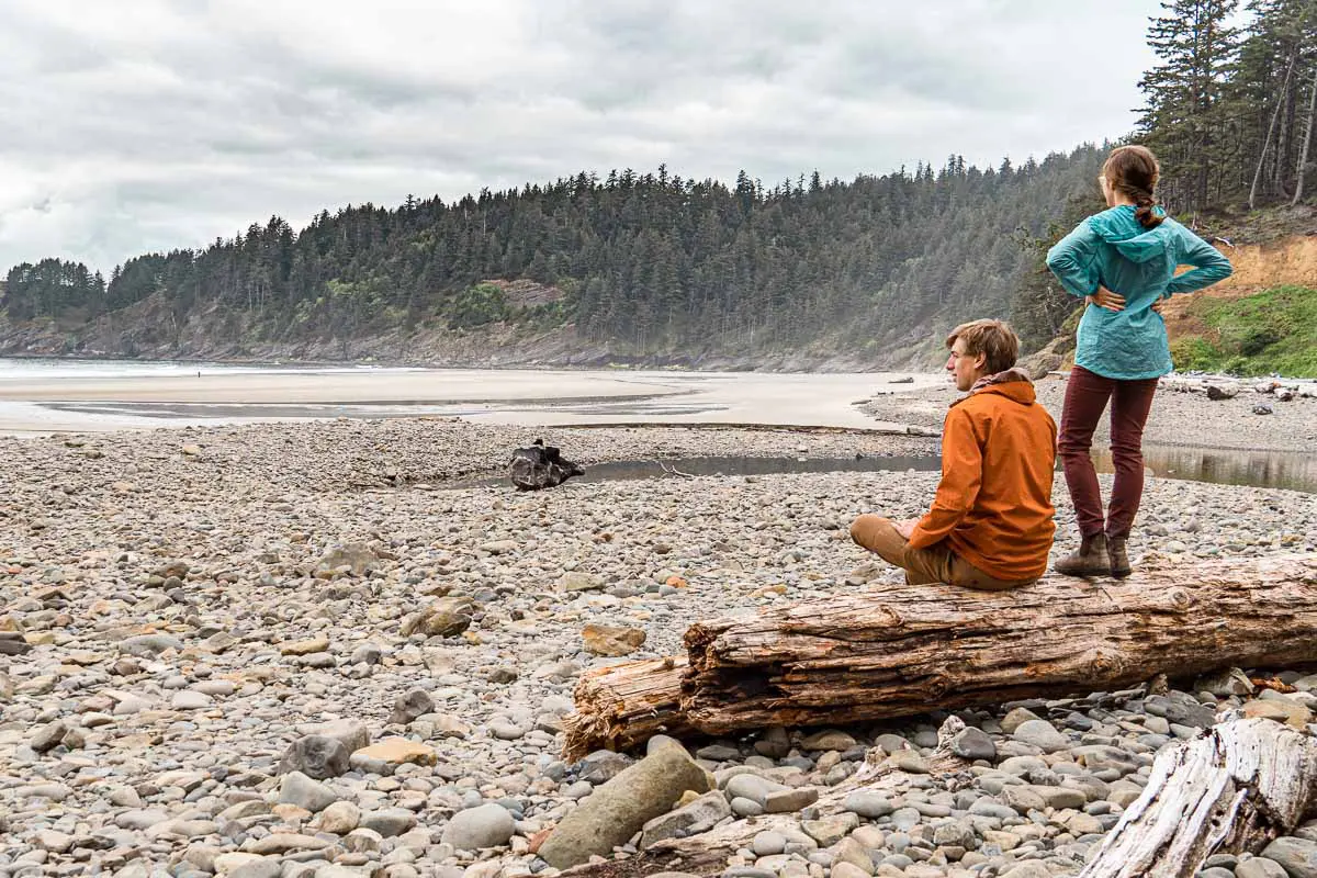 Michael is sitting on a log and Megan stands on the log next to him. They are looking out at a rocky beach.